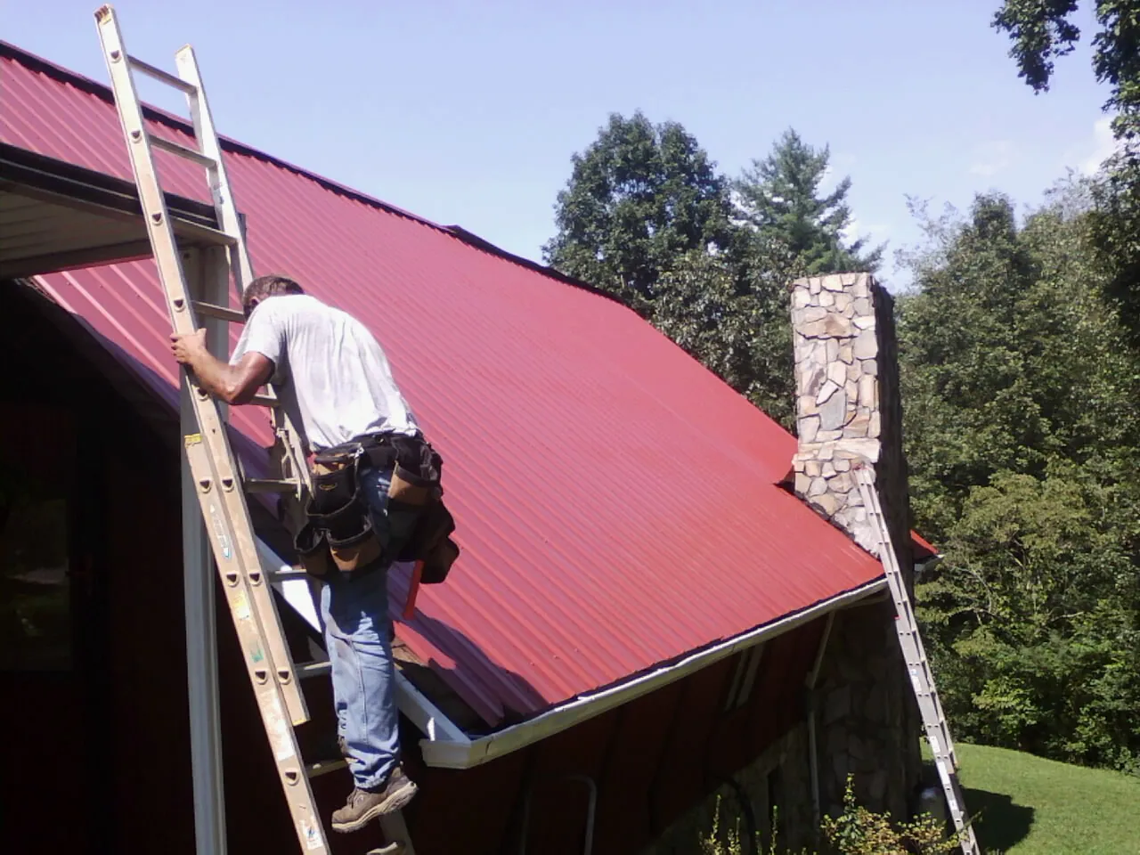 Roofer on a ladder beside a red metal roof and stone chimney — Soffit & Fascia Repair by Roofing Brothers in San Martin